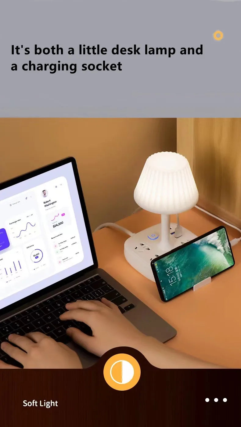 A laptop is open on a desk next to a white desk lamp and a smartphone charging on the surface.