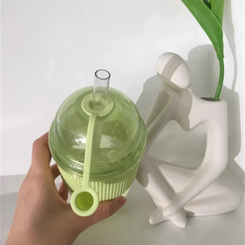 Eine Hand hält ein hellgrünes Glas mit Strohstopfen neben einem weißen Dekorationsgegenstand und einer grünen Pflanze.