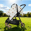 A white stroller cover with gold stars is shown on a black stroller frame outdoors in a grassy field.