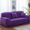 A deep purple elastic stretch sofa cover with two accent pillows placed on a wooden floor near a bright window.