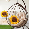 A beige floor pillow shaped like a sunflower is nestled inside a large, dark metal hanging chair.