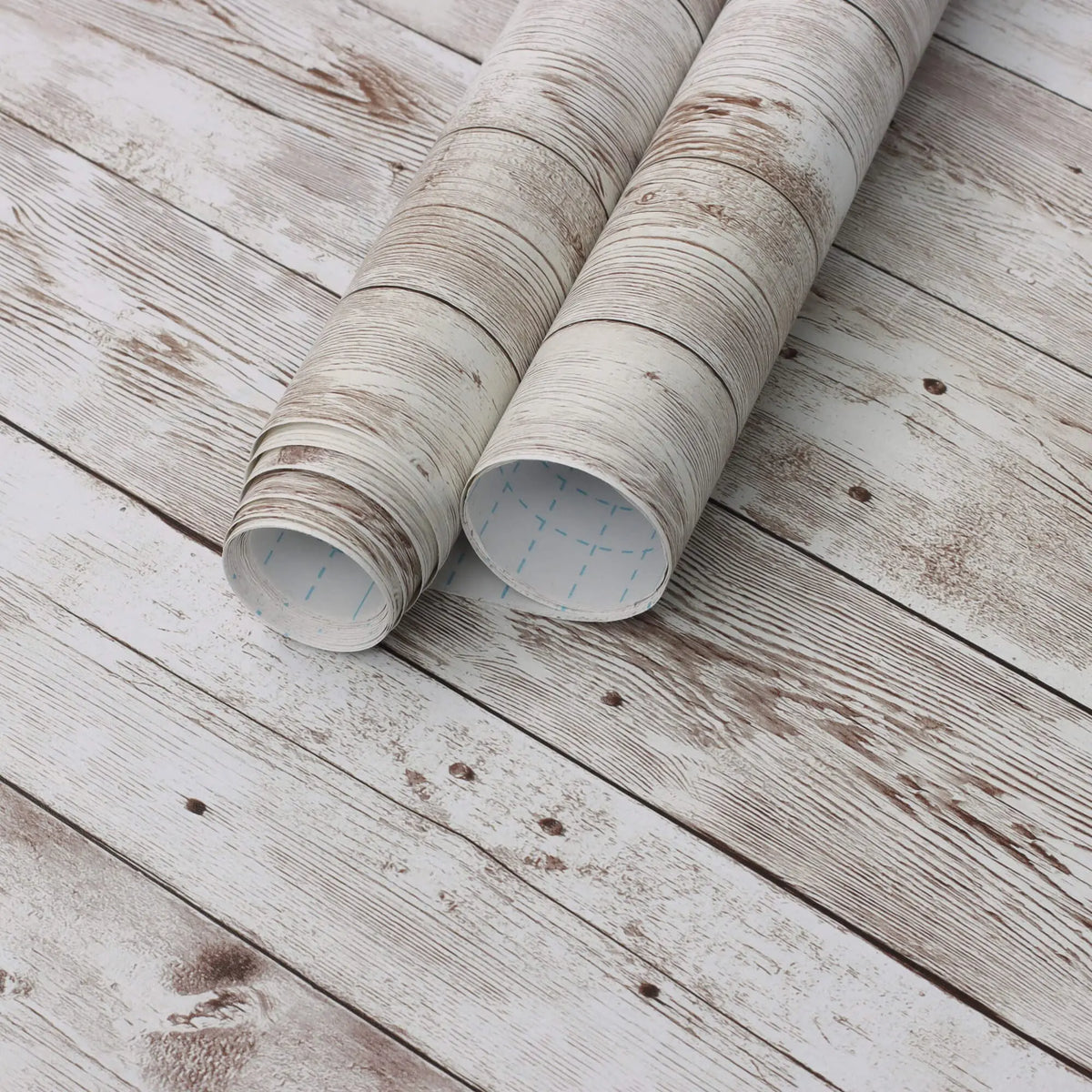 Rolls of retro wood grain self-adhesive wallpaper lying on a weathered white wooden surface.