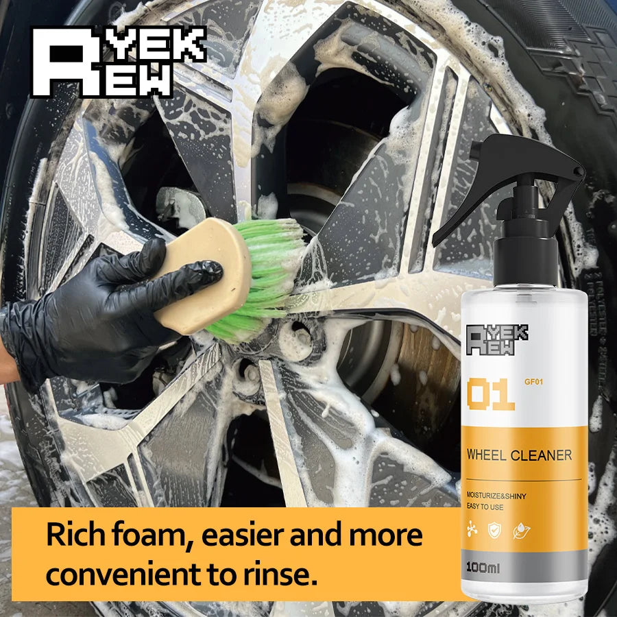 A hand is cleaning a car wheel with a yellow brush and a spray bottle of wheel cleaner foam.