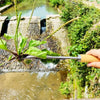A hand holds a carbon steel manual weeding tool set with a sickle, weeder, and hand shovel near a small water feature.