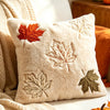 A cream-colored plush throw pillow featuring embroidered maple leaves in shades of orange, green, and brown.