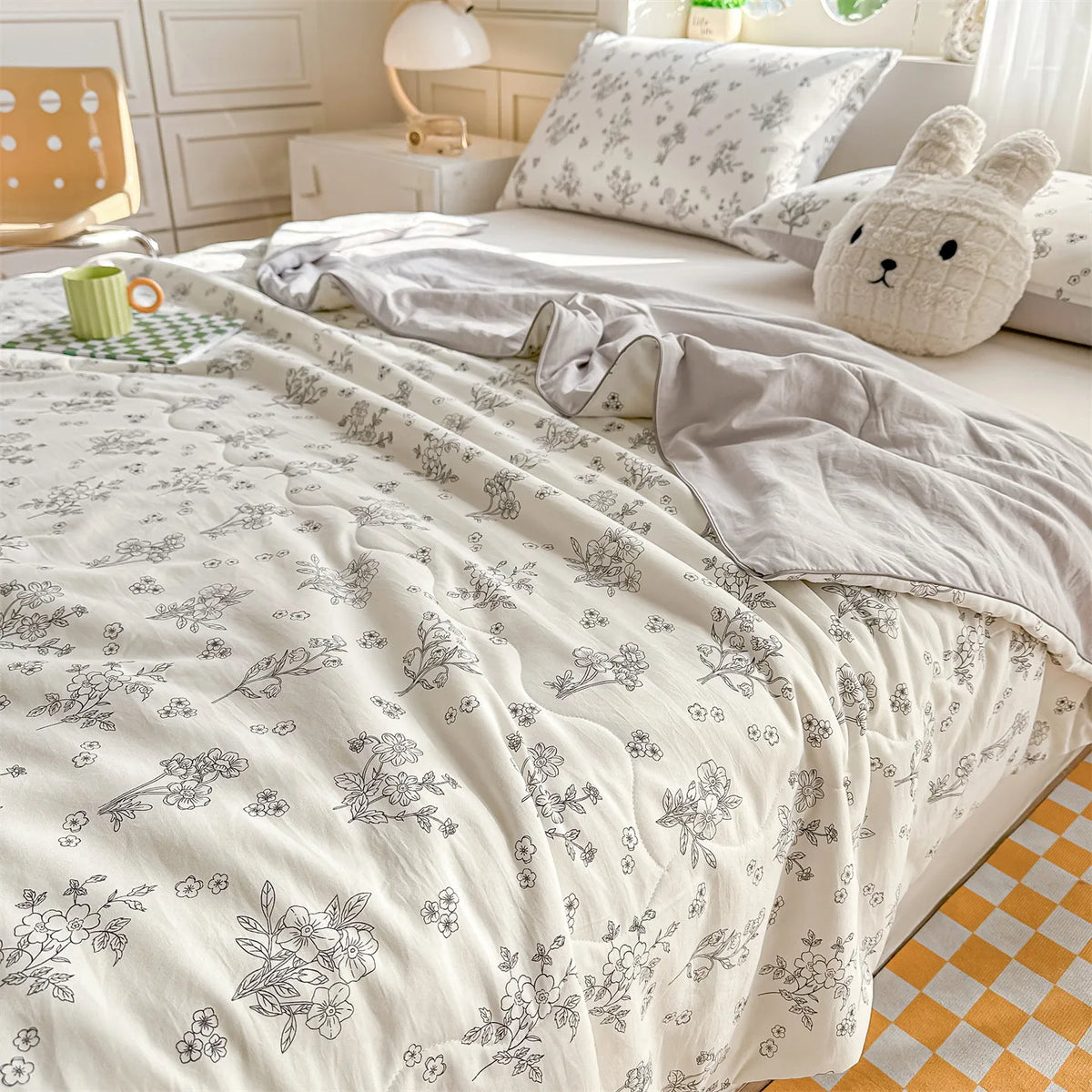 A white quilt with a delicate floral print is displayed on a bed, next to a cream-colored stuffed animal.