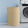 A cylindrical striped laundry basket in beige and white with gold handles next to a white washing machine.