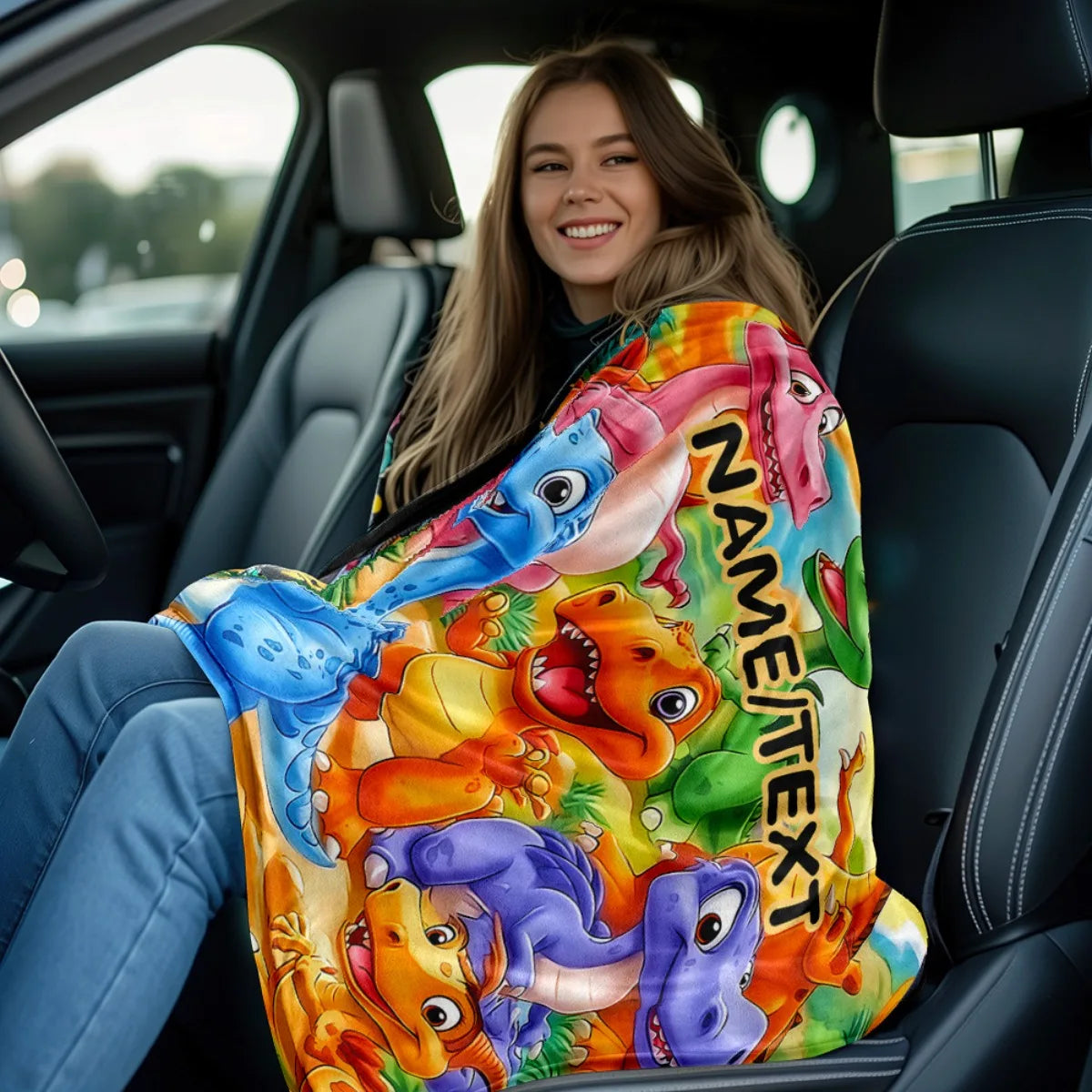 A young woman is sitting in a car, holding a colorful personalized dinosaur blanket with various dinosaurs and text on it.