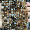 A close-up of natural stone square faceted beads in various shades of brown, amber, and black stacked on a hand.