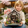 A young boy holds a detailed miniature Japanese sakura tea house kit featuring pink blossoms and wooden elements.