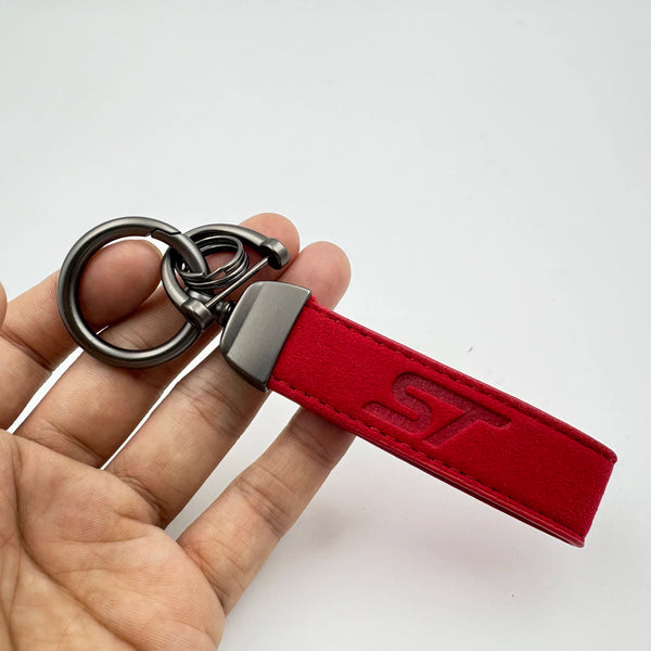 A hand holds a red suede leather keychain with a silver split ring and an ST logo stamp.