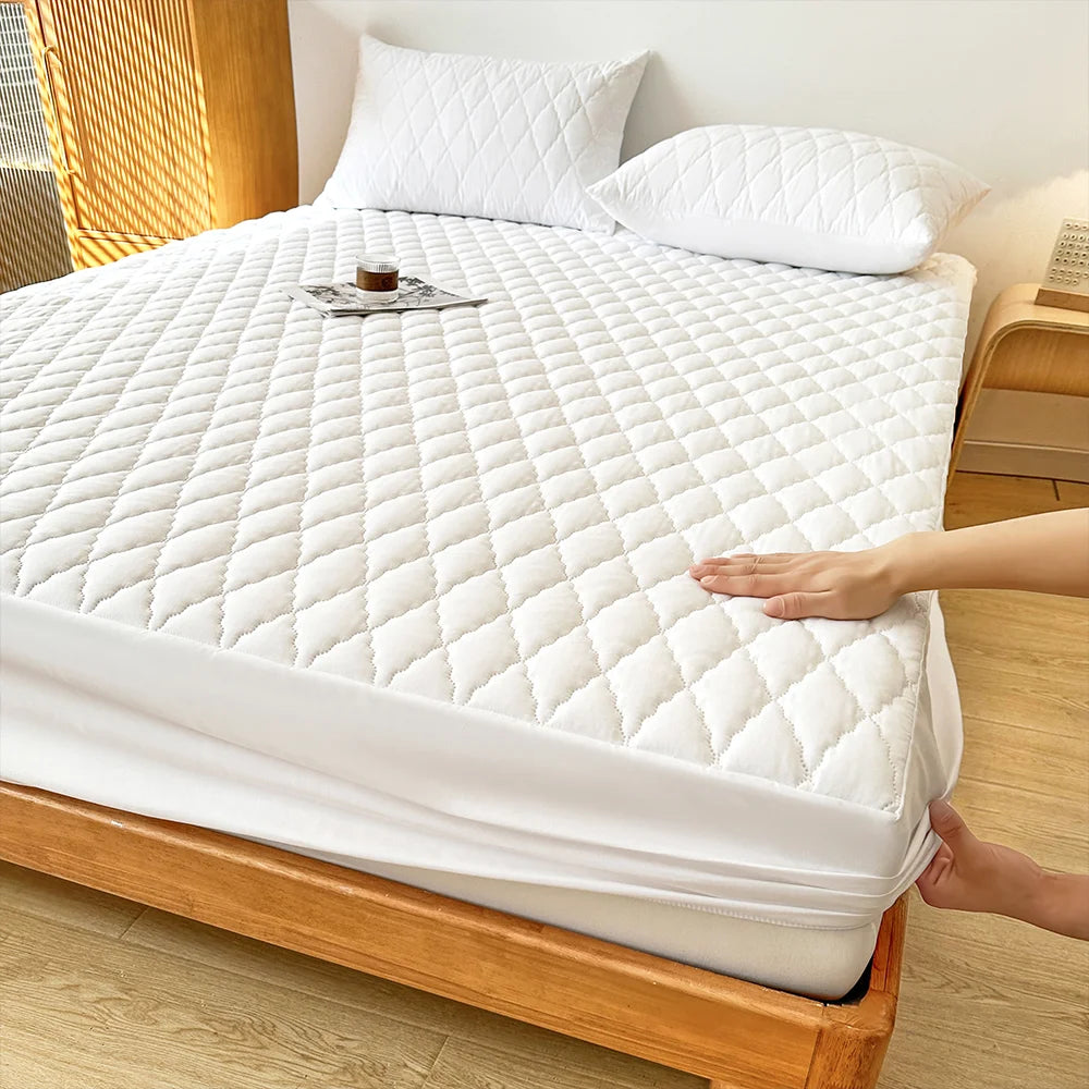 A white waterproof fitted mattress protector is shown on a wooden bed frame, with a hand resting on the fabric.