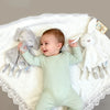 A baby wearing a light green onesie is surrounded by a white blanket and a gray elephant plush toy with a teether.