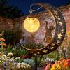 A solar owl garden light with moon decor illuminates a night garden setting against a brick wall.