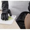 A hand wearing black rubber cleaning gloves is handling a piece of green vegetable on a white cutting board.