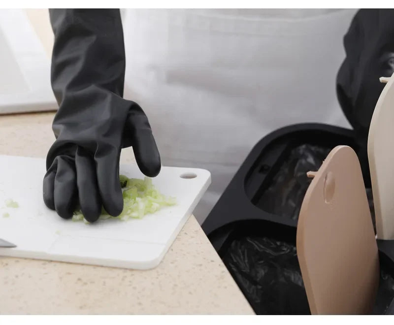 A hand wearing black rubber cleaning gloves is handling a piece of green vegetable on a white cutting board.