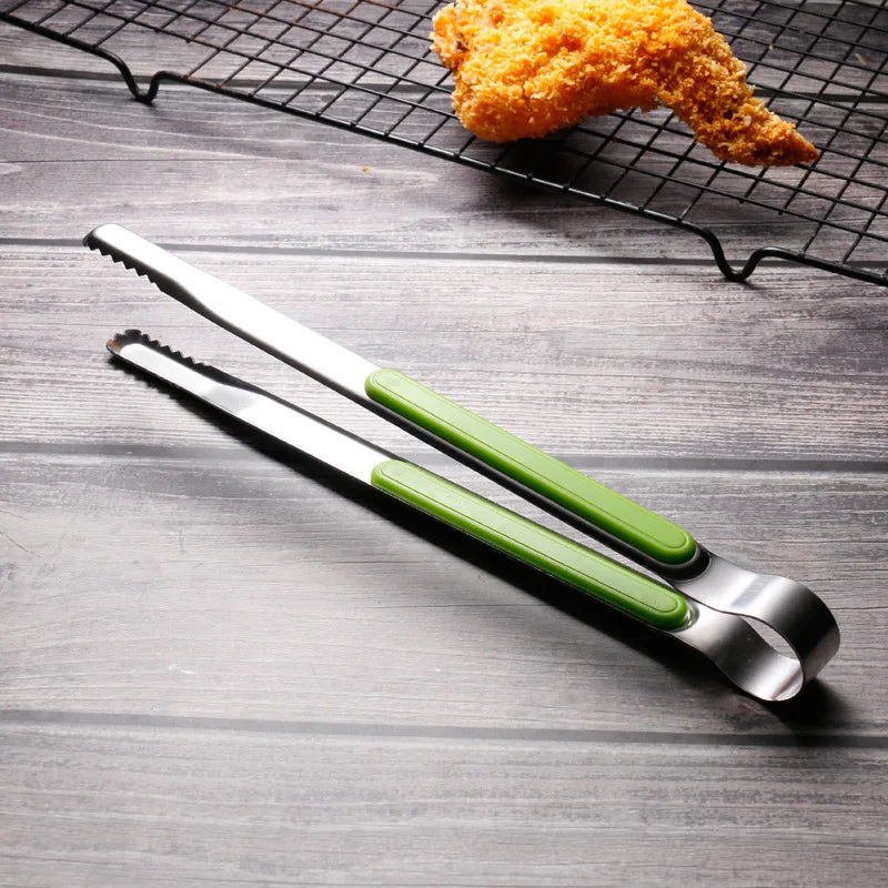 A pair of stainless steel BBQ tongs with green handles resting on a dark wooden surface next to a piece of fried food on a wi