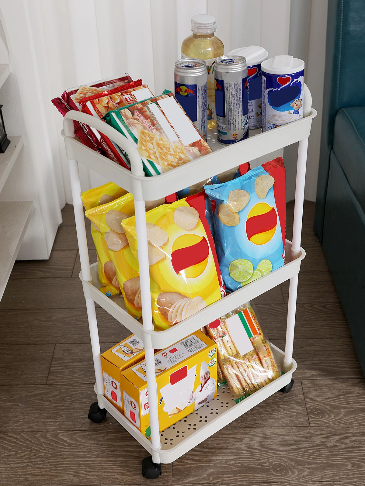 A white 3-tier plastic utility cart filled with various snack foods and drinks.
