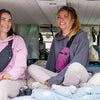 Two women sitting inside a van looking at the camera with pillows and blankets.