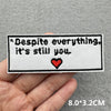 Black embroidered patch with the text Despite everything it's still you and a red heart on a gray fabric background