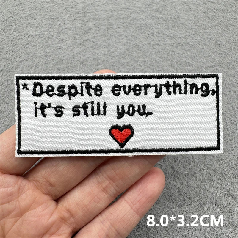 Black embroidered patch with the text Despite everything it's still you and a red heart on a gray fabric background