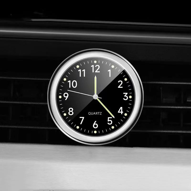 A close-up of a round quartz clock with luminous hands mounted on a black car vent grille.