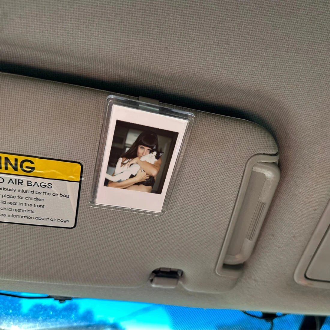 A vertical photo frame clip is mounted on a car visor, next to a yellow warning sign about air bags.