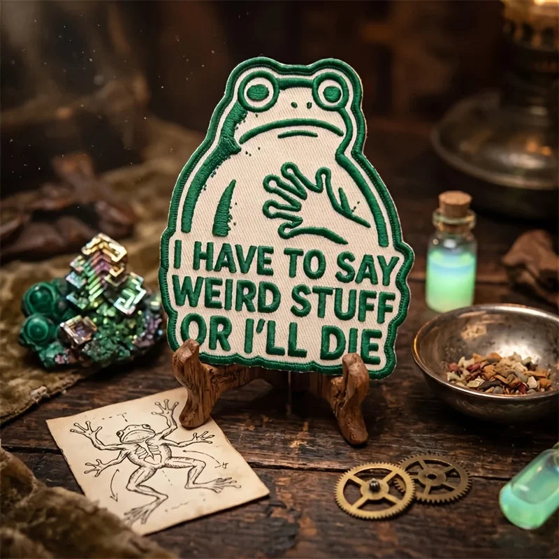 An embroidered patch of a frog saying I have to say weird stuff or I'll die resting on wood next to crystals and gears.