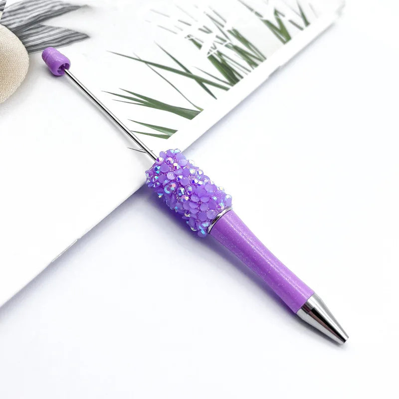 A purple ballpoint pen with a sparkly purple sequin tip resting on a white background next to green foliage.