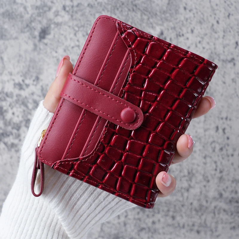 A hand holds a deep burgundy crocodile-embossed clutch wallet with a pink leather strap.