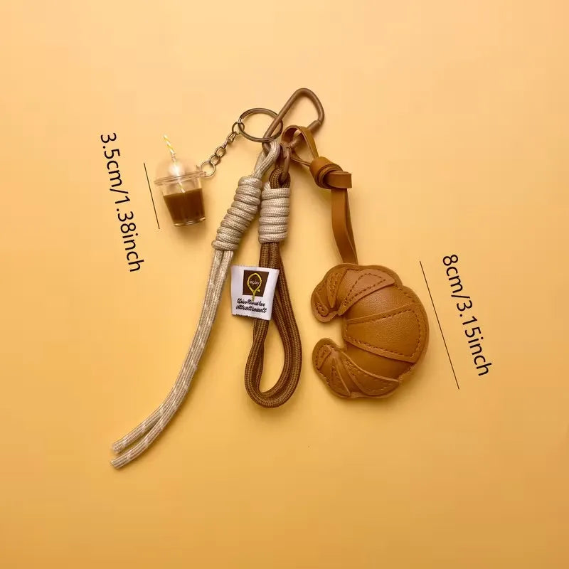 A braided leather croissant keychain with a bag pendant displayed on a beige background.