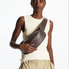 Model in cream sleeveless top and tan pants holding a dark brown genuine leather crossbody bag.