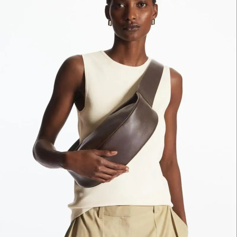 Model in cream sleeveless top and tan pants holding a dark brown genuine leather crossbody bag.