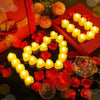 A display of heart-shaped yellow LED candles surrounded by red rose petals on a dark surface.