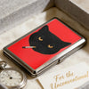 A vintage metal card and cash case featuring a black cat silhouette on a red background resting on a light fabric.
