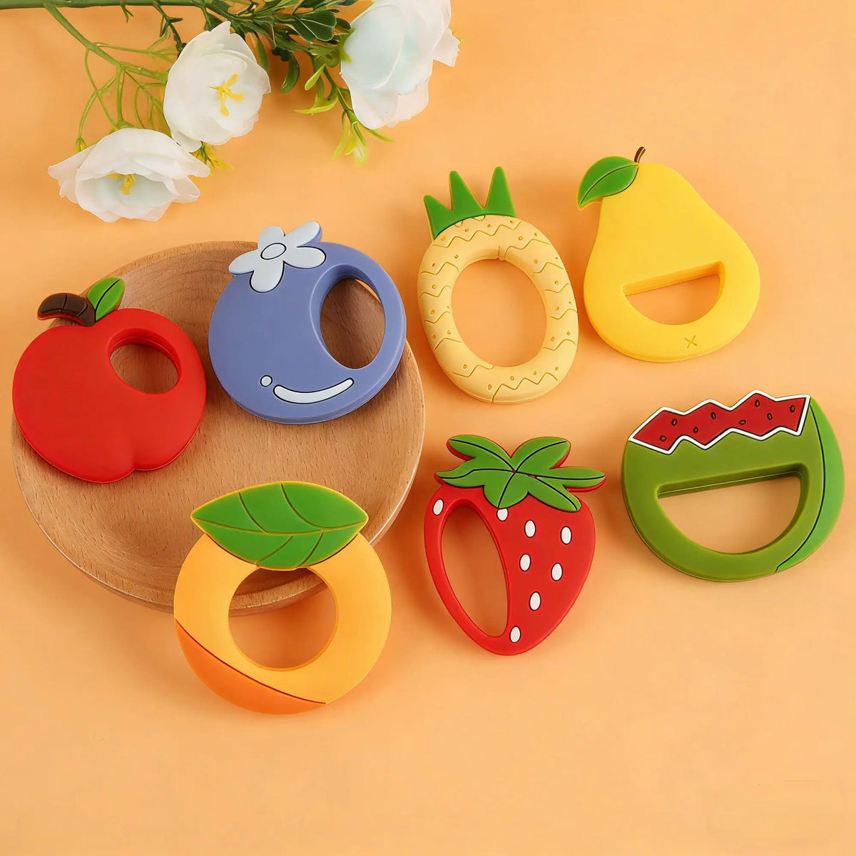 A collection of silicone fruit-shaped teethers including an apple, blueberry, lemon, and strawberry, arranged on a light back