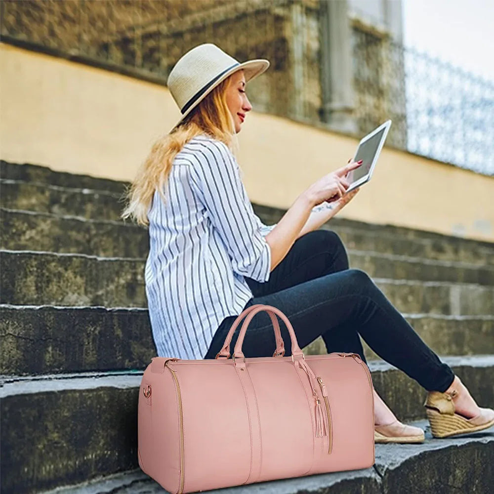 A woman wearing a hat is sitting on stone steps, looking at a tablet while holding a pink foldable travel duffel bag.