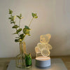 A gold-framed glass vase holds green foliage next to a clear acrylic bear night light.