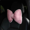 A pink silky bow shaped headrest pillow with a white trim is placed on a black car seat.