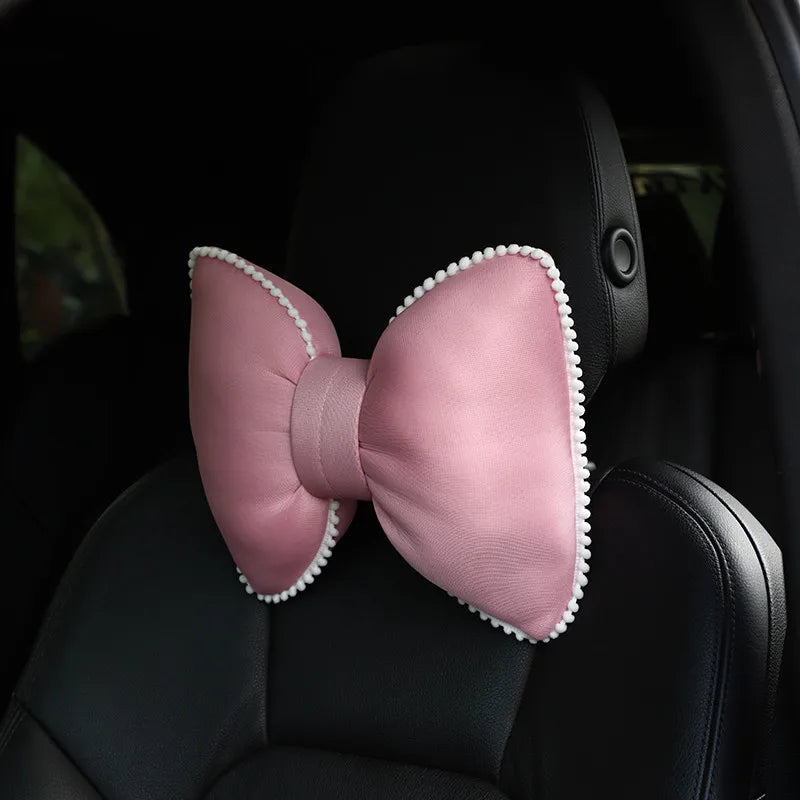 A pink silky bow shaped headrest pillow with a white trim is placed on a black car seat.