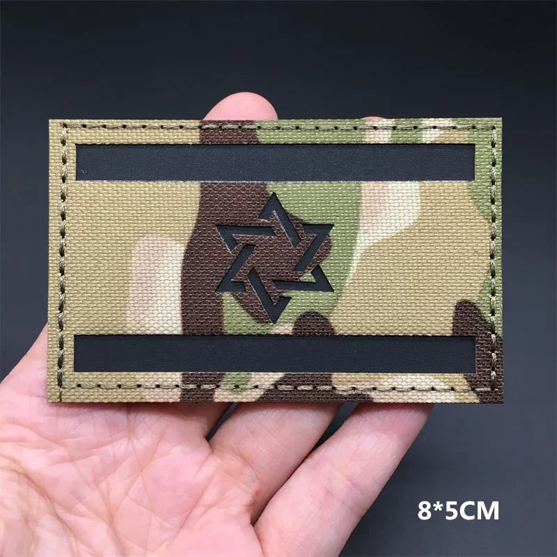 A rectangular tactical patch featuring a camouflage pattern with a central Star of David symbol on a khaki background.