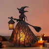 A dark metal witch figure with a pointed hat and broom, illuminating a glowing lantern on a wooden surface.