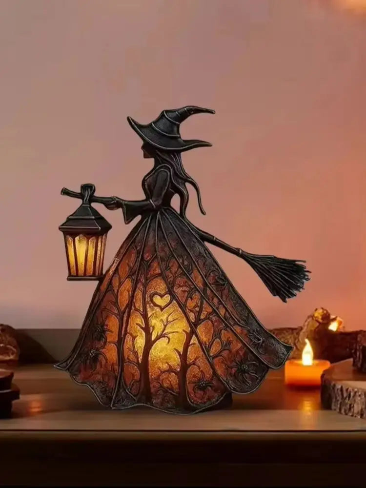 A dark metal witch figure with a pointed hat and broom, illuminating a glowing lantern on a wooden surface.