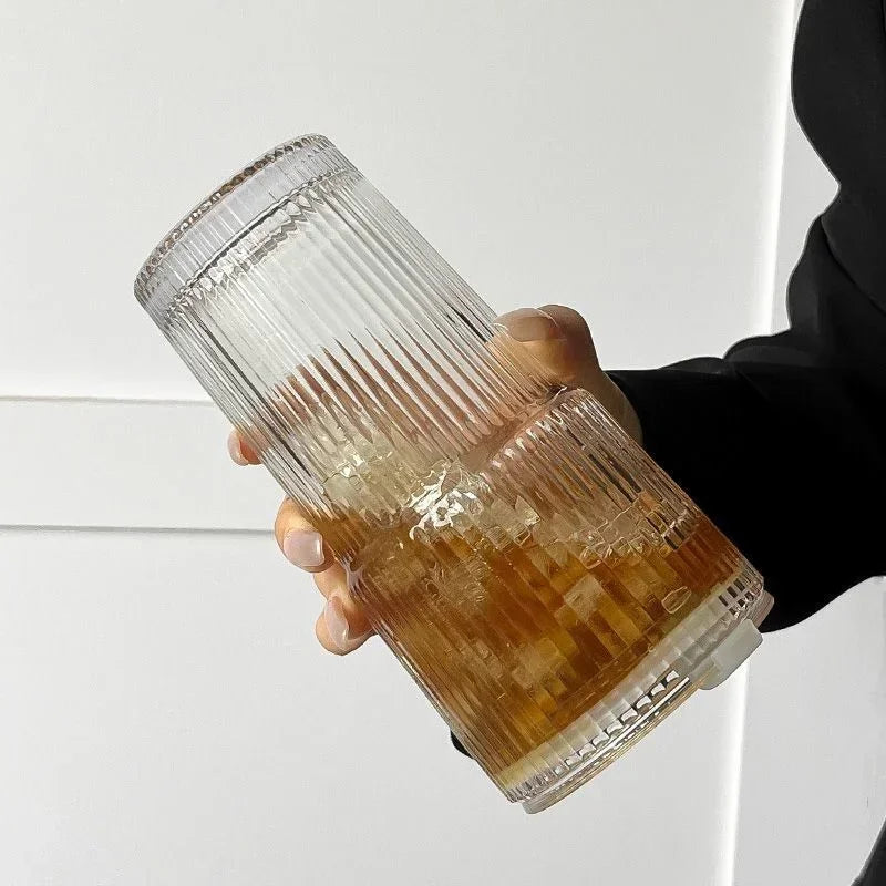 Une main tient un verre tumbler rayé de 600ml avec couvercle et paille, montrant sa texture de verre ambré clair.