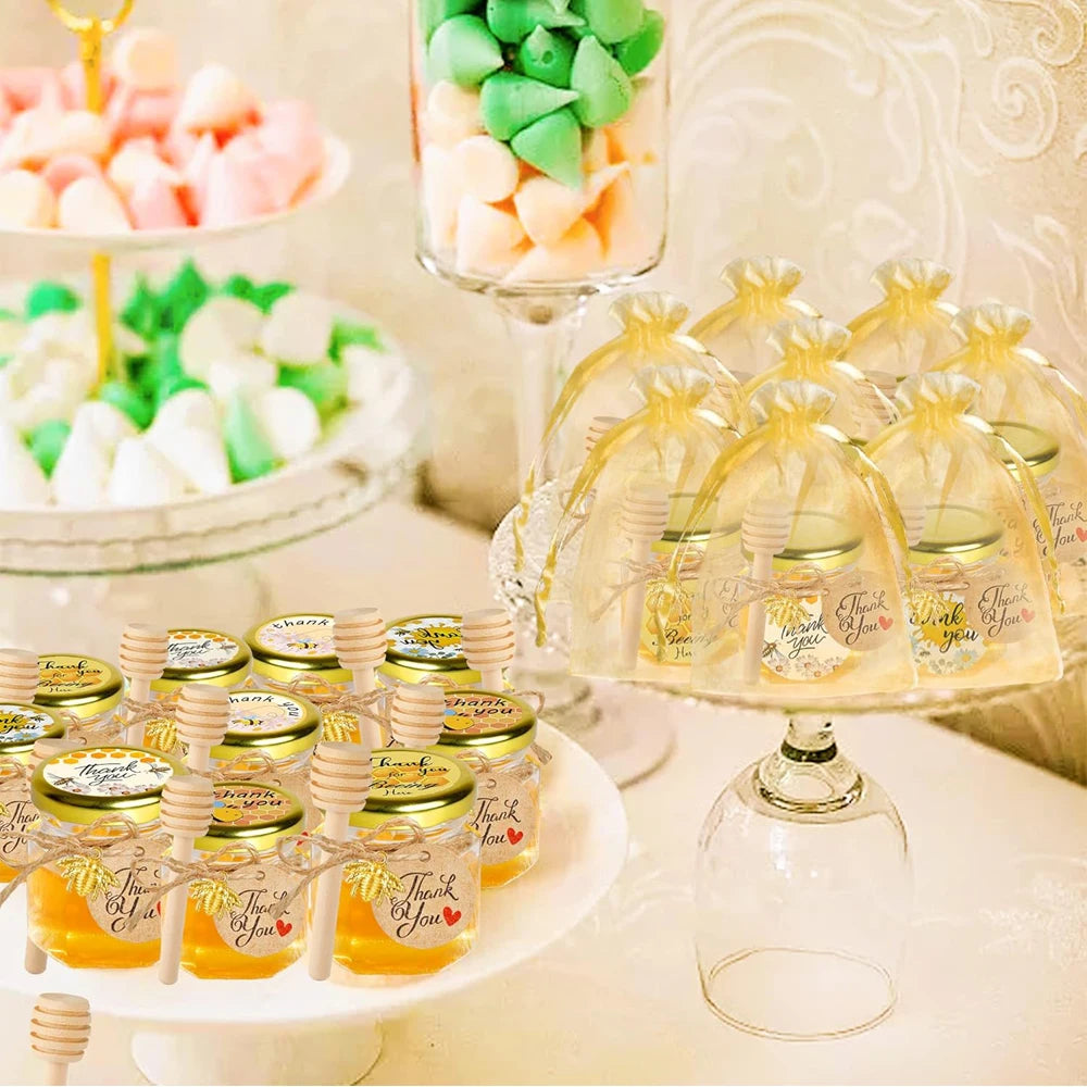 A display of hexagon glass honey jars with wood dippers and bee pendants on a white plate.