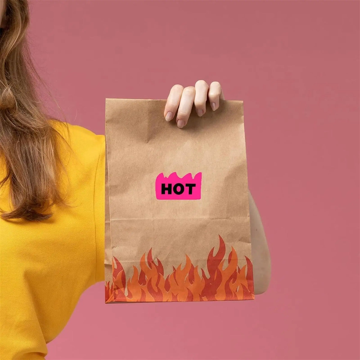 A hand is holding a brown paper bag with a pink 'HOT' sticker on it against a pink background.