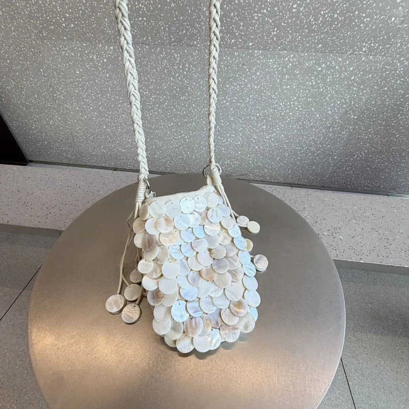 A white sequin accent chain shoulder bag with a woven strap displayed on a round pedestal.