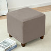 A square, taupe fabric ottoman slipcover with a textured diamond pattern resting on a light carpet.