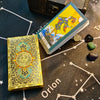 A gold foil tarot card deck and guidebook are displayed on a dark surface with Orion constellation markings.