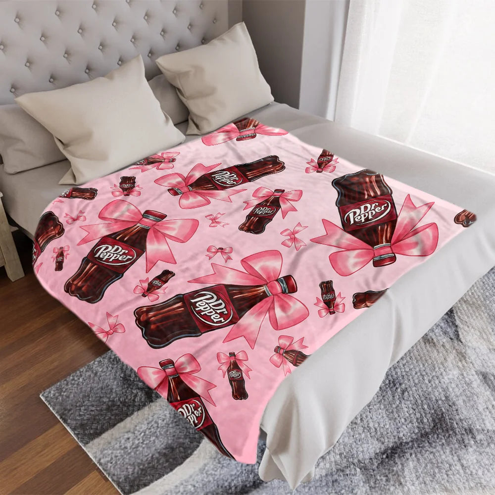 A pink Dr. Pepper ultra-soft printed throw blanket is draped over a white bed with a light gray textured throw.